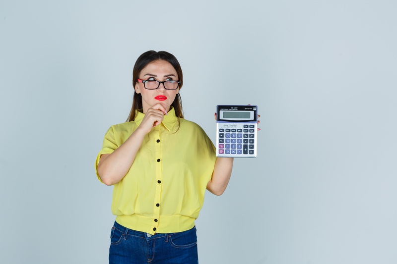 Mujer con camisa amarilla sosteniendo una calculadora grande y mirando pensativa, representando la importancia de analizar el presupuesto personal y controlar los gastos